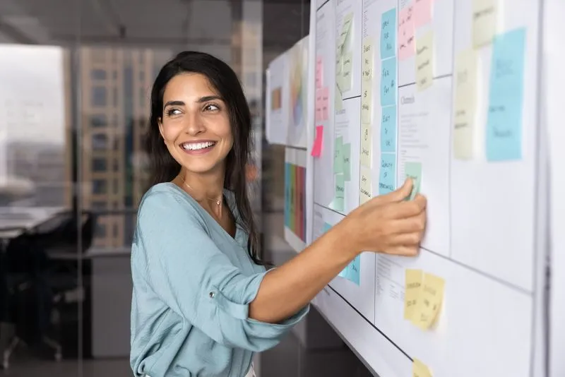 Junge Frau benutzt einen Klebezettel um etwas auf ein gut befülltes Whiteboard zu heften